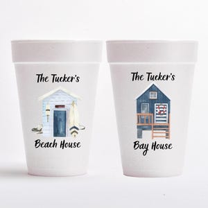 May include: Two white foam cups with beach house illustrations. One cup features a light blue beach house with the text "Beach House" and "The Tucker's". The other cup has a dark blue bay house with the text "Bay House" and "The Tucker's".