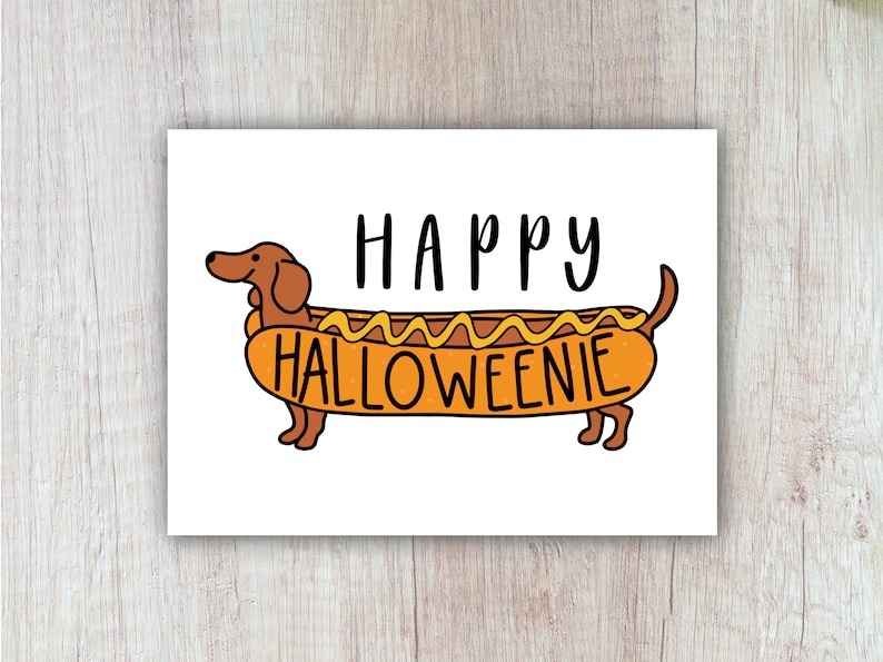 May include: A rectangular greeting card featuring a cartoon illustration of a brown dachshund dog inside a hot dog bun with yellow mustard on top. The text HAPPY HALLOWEENIE is printed in black, with HAPPY above the dog and HALLOWEENIE on the bun.