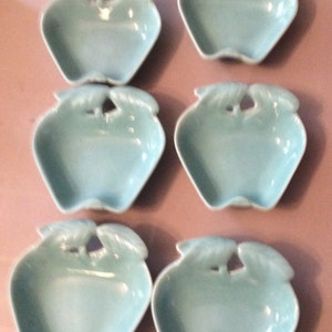 May include: Six light blue ceramic apple-shaped dishes. Each dish has a stem and leaf design at the top and a small opening in the center. The dishes are arranged in two rows of three.