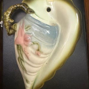 May include: A vintage ceramic wall pocket in the shape of a leaf, with a handle and a hole for hanging. The pocket is painted in shades of green, cream, and pink, with a floral design. A decorative piece for the home.
