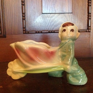 Op de afbeelding: Een vintage keramieken bloempot in de vorm van een zittende figuur, met een lichtgroen outfit en bruin haar. De bloempot heeft een bloemvormige opening met roze en witte accenten. De figuur rust op een groene basis.