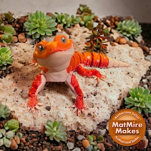 May include: A 3D printed orange and white gecko figurine with red spots. The gecko is posed on a rock with green succulents in the background. The image includes a circular sticker with the text "Authorized Seller MatMire Makes Authorized Seller".