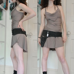 May include: A woman wearing a gray sleeveless top with a black belt and black combat boots. The top has a cowl neck and the belt has a buckle and a silver charm.