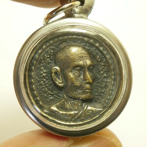 May include: A silver pendant with a round, bronze medallion featuring a detailed profile of a monk. The medallion is surrounded by a decorative border.