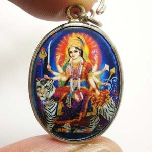 Puede incluir: Un colgante ovalado de plata con una imagen colorida de la diosa hindú Durga montando un tigre blanco. La imagen está rodeada por un borde azul.