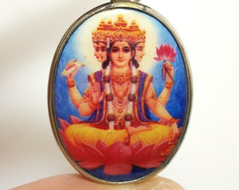 May include: Oval-shaped silver pendant with a colorful image of a four-armed Hindu deity sitting on a lotus flower. The deity has three faces and is wearing a crown and jewelry.