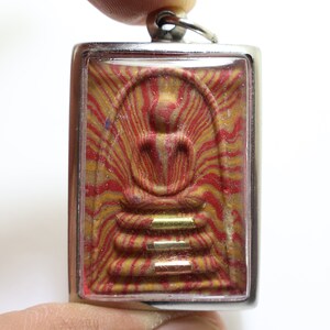 Könnte beinhalten: Ein silberner Anhänger mit einem roten und goldenen Bild eines Buddha im Inneren. Der Anhänger ist in einer durchsichtigen Plastikhülle eingeschlossen.