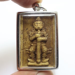 Puede incluir: Un colgante de plata con un frente de vidrio transparente, que muestra una talla detallada de una figura de pie de color marrón claro. La figura lleva una corona y sostiene una espada.