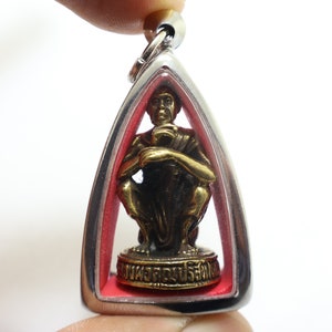 May include: Silver pendant with a small gold statue of a seated Buddha inside. The pendant has a red background and the words "พระพุทธเจ้า" are inscribed on the base of the statue.