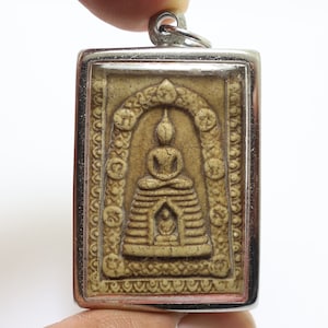 Könnte beinhalten: Ein silberner Anhänger mit einem beigefarbenen Buddha-Amulett. Das Amulett ist rechteckig und zeigt eine detaillierte Schnitzerei eines sitzenden Buddha in einem dekorativen Bogen.