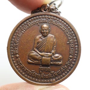 Könnte beinhalten: Ein bronzefarbener Anhänger mit einer sitzenden Figur in traditioneller Kleidung. Der Anhänger hat eine runde Form mit geprägtem Text am Rand und einer kleinen Schlaufe zur Befestigung. Die Figur ist zentriert.