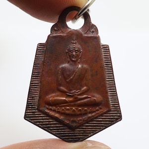 Könnte beinhalten: Kupferfarbener Anhänger mit einer sitzenden Buddha-Figur in meditativer Haltung. Der Anhänger hat eine rechteckige Form mit dekorativem Rand und einer Schlaufe zum Aufhängen. Der Buddha ist mit detaillierten Gesichtszügen und drapierten Gewändern dargestellt.