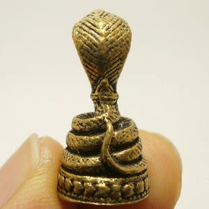 Puede incluir: Una figurita de metal de color dorado que representa una cobra con la cabeza levantada y el cuerpo enroscado en espiral. La figurita est&aacute; detallada con escamas y tiene una base decorativa.