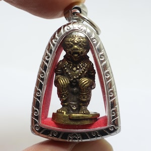 Puede incluir: Una pequeña figura de color bronce de una deidad sentada, encerrada en un colgante plateado en forma de arco. La figura está detallada con intrincados patrones y descansa sobre una base roja. El colgante tiene un lazo para colgar.