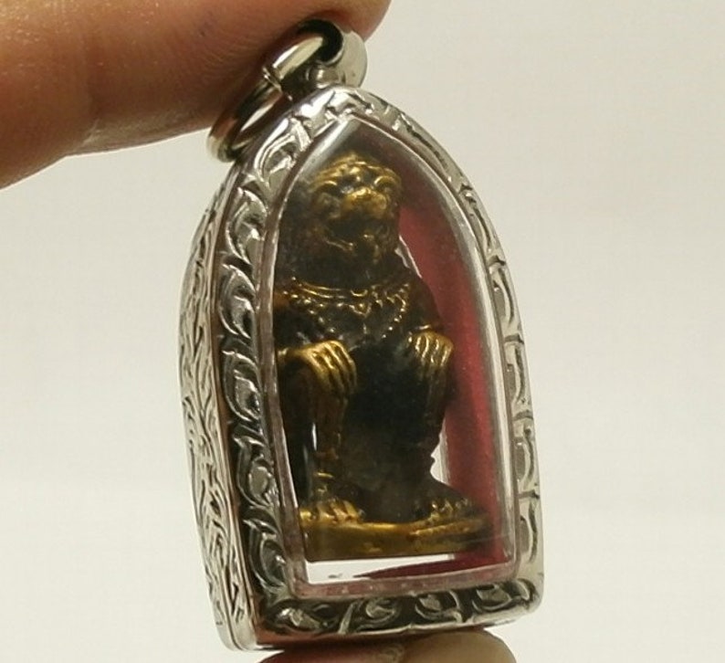 Pu&ograve; includere: Un ciondolo d'argento con un inserto di vetro trasparente che contiene una piccola statua dorata di una scimmia. Il ciondolo presenta dettagli in argento intricati attorno al bordo.