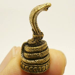 Puede incluir: Una peque&ntilde;a figura de metal dorada de una cobra con la cabeza levantada y la capucha expandida. La serpiente est&aacute; enroscada sobre s&iacute; misma y tiene una superficie detallada y texturizada.