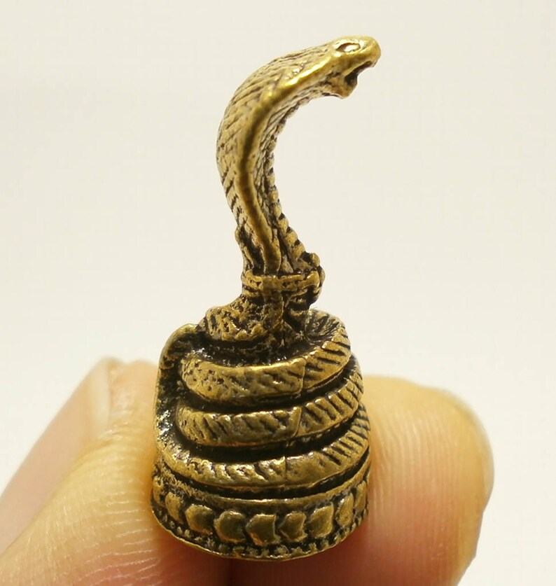 Puede incluir: Una peque&ntilde;a figura de metal dorada de una cobra con la cabeza levantada y la capucha expandida. La serpiente est&aacute; enroscada sobre s&iacute; misma y tiene una superficie detallada y texturizada.