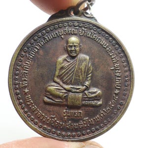 Könnte beinhalten: Eine runde Bronzemedaille mit einem erhabenen Bild eines sitzenden Mönchs. Die Medaille hat einen kreisförmigen Rand mit einem sich wiederholenden Muster aus kleinen Kreisen. Der Text am Rand der Medaille lautet "พระครูวิสุทธิ์ธรรมรัตน์  วัดไตรมิตรวิทยารามวรวิหาร  กรุงเทพมหานคร".