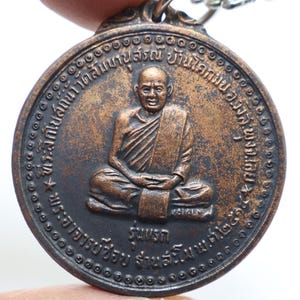 Könnte beinhalten: Ein runder Anhänger aus Bronze mit einem erhabenen Bild eines Mönchs in sitzender Meditationshaltung. Der Anhänger hat einen kreisförmigen Rand mit einem sich wiederholenden Muster aus kleinen Kreisen. Der Text um den Rand lautet "พระอาจารย์ฝั้น อาจาโร วัดป่าอุดมสมพร"