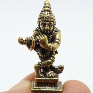 Könnte beinhalten: Kleine, detaillierte Messingfigur einer Gottheit, die Flöte spielt. Die Figur ist in einer dynamischen Pose dargestellt, mit komplizierten Details in Kleidung und Kopfschmuck. Die Statue ist auf einem quadratischen Sockel montiert.