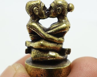Puede incluir: Pequeña escultura de color bronce que representa dos figuras abrazadas, con sus rostros cerca en un beso. Las figuras están sentadas, con las piernas entrelazadas. La escultura está montada sobre una base circular.
