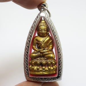 May include: A silver pendant with a gold-colored Buddha statue inside. The pendant is shaped like a triangle with a decorative border.
