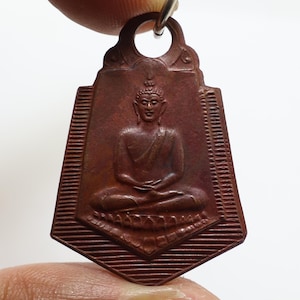 Könnte beinhalten: Ein rotbrauner Anhänger mit einer sitzenden Buddha-Figur in meditativer Haltung. Der Anhänger hat eine rechteckige Form mit dekorativer Oberseite und einem Ring zum Aufhängen. Der Buddha ist mit detaillierten Gesichtszügen und Gewändern dargestellt.
