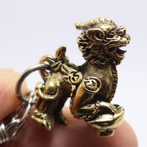 May include: A gold-colored metal keychain featuring a detailed depiction of a mythical lion-like creature with wings. The creature is sitting on its haunches with its head turned to the side, and its mouth is slightly open. The keychain is attached to a silver-colored metal ring.