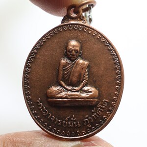 Könnte beinhalten: Ein kupferfarbener Anhänger mit einer sitzenden Figur eines Mönchs mit einem detaillierten Gesicht und einem gemusterten Rand. Der Anhänger hat thailändischen Text am Rand, der "พระอาจารย์ฝั้น อาจาโร" lautet.
