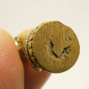 Puede incluir: Primer plano de una figurita de metal de color dorado con una base circular. La base tiene un borde decorativo y un dise&ntilde;o en relieve en el centro.