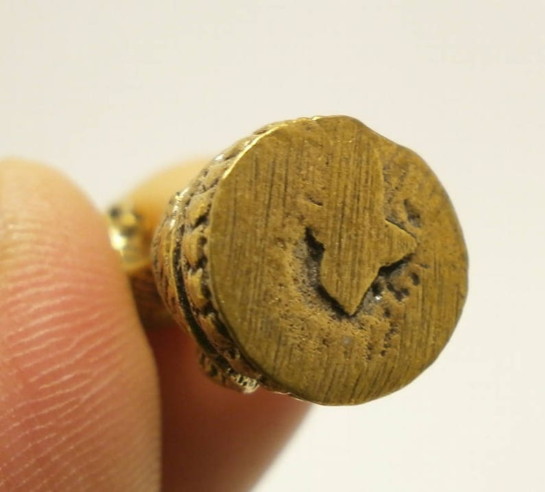 Puede incluir: Primer plano de una figurita de metal de color dorado con una base circular. La base tiene un borde decorativo y un dise&ntilde;o en relieve en el centro.