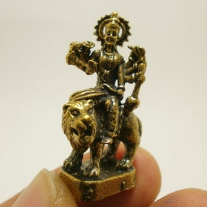 Pode incluir: Uma figurinha de metal dourada de uma divindade a montar num leão. A divindade tem múltiplos braços e usa uma coroa. A figurinha está sobre uma base quadrada.
