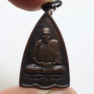 Puede incluir: Colgante de bronce con una parte superior triangular y una figura sentada con túnicas. La figura tiene una expresión serena y el colgante tiene un aspecto oscuro y envejecido. Hay un pequeño lazo en la parte superior para sujetarlo.