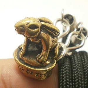 Può includere: Un ciondolo a forma di coniglio dorato con un designo dettagliato, seduto su una base rotonda con caratteri thailandesi. Il ciondolo è attaccato a una catena d'argento.