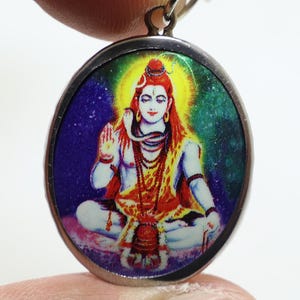May include: A circular, silver-toned pendant displaying a vibrant image of a deity with a yellow aura, red hair, and multiple necklaces. The background is a blend of blue, green, and purple. The pendant is suspended by a silver chain.