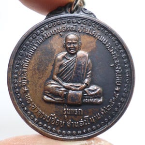 Könnte beinhalten: Eine runde Bronzemedaille mit einem erhabenen Bild eines Mönchs in sitzender Meditationshaltung. Die Medaille hat einen thailändischen Text am Rand.