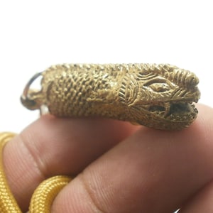 Può includere: Un ciondolo dorato a forma di serpente con una superficie dettagliata e testurizzata. La testa del serpente è sollevata e ha un'espressione minacciosa con denti affilati.