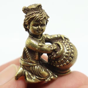 Puede incluir: Pequeña y detallada figura de latón de un niño sentado sosteniendo un recipiente redondo y estampado. El niño tiene el pelo rizado y lleva una prenda drapeada. La figura tiene un aspecto vintage.