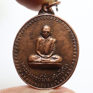 Könnte beinhalten: Ein kupferfarbener Anhänger mit einem erhabenen Bild eines Mönchs in sitzender Meditationshaltung. Der Anhänger ist oval und hat einen dekorativen Rand. Der Text "พระอาจารย์  กว้าน" ist um das Bild herum eingeschrieben.