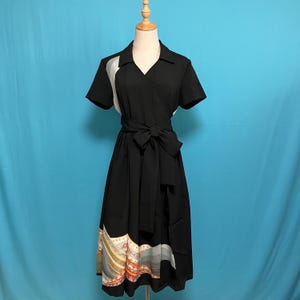May include: A black wrap dress with short sleeves and a V-neck. The dress features a contrasting white panel on the left shoulder and a patterned hemline with orange, gold, and gray accents. A matching black belt cinches the waist.