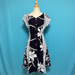 以下が含まれることがあります： 白のフローラルと幾何学模様のネイビーブルーのドレス。 ドレスはVネック、半袖、フロントボタン留めが特徴です。 ドレスは青い背景のマネキンに展示されています。