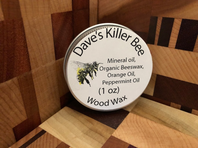 May include: A white round tin with a black and white label that says "Dave's Killer Bee Wood Wax (1 oz)". The label also lists the ingredients: mineral oil, organic beeswax, orange oil, and peppermint oil. A cartoon bee is pictured on the label.