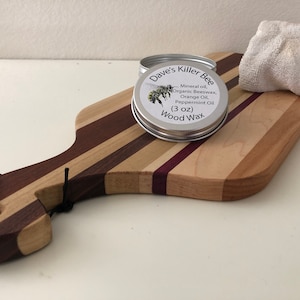 May include: A silver tin of Dave's Killer Bee Wood Wax, with a bee illustration on the label. The label reads "Mineral oil, Organic Beeswax, Orange Oil, Peppermint Oil (3 oz) Wood Wax". The tin is sitting on a wooden cutting board with a white cloth in the background.