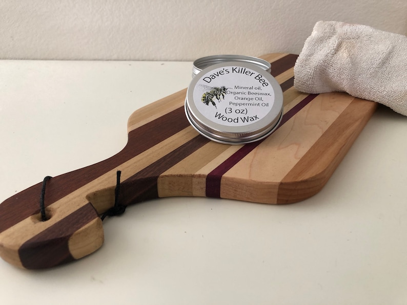 May include: A silver tin of Dave's Killer Bee Wood Wax, with a bee illustration on the label. The label reads "Mineral oil, Organic Beeswax, Orange Oil, Peppermint Oil (3 oz) Wood Wax". The tin is sitting on a wooden cutting board with a white cloth in the background.