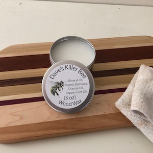 May include: A silver tin of Dave's Killer Bee Wood Wax, a 3 ounce container of wood polish with a bee illustration on the label. The label reads "Mineral oil, Organic Beeswax, Orange Oil, Peppermint Oil". The tin is sitting on a wooden cutting board with a white cloth next to it.