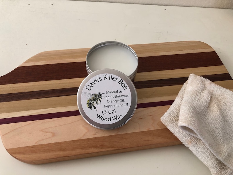 May include: A silver tin of Dave's Killer Bee Wood Wax, a 3 ounce container of wood polish with a bee illustration on the label. The label reads "Mineral oil, Organic Beeswax, Orange Oil, Peppermint Oil". The tin is sitting on a wooden cutting board with a white cloth next to it.