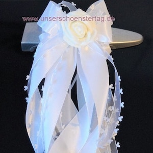 Puede incluir: Decoración de boda blanca con un gran lazo y largas cintas fluidas. Una rosa color crema está en el centro del lazo. Las cintas están adornadas con pequeños detalles florales blancos. El texto "www.unserschoensterTag.de" es visible.