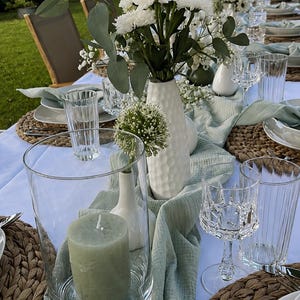 Peut inclure: Une table dressée avec un chemin de table en gaze vert sauge, des sets de table ronds tressés, des vases en céramique blanche avec des fleurs blanches, des verres transparents et une bougie pilier vert sauge dans un vase cylindrique en verre.