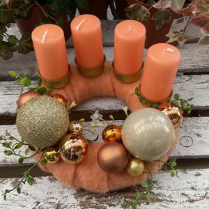 Könnte beinhalten: Ein orangefarbener Adventskranz mit vier orangenen Kerzen und goldenen Akzenten. Der Kranz ist mit goldenen, braunen und weißen Kugeln verziert.