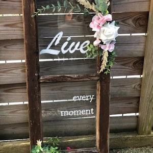 Könnte beinhalten: Ein rustikaler Holzfensterrahmen mit einer Glasscheibe. Die Glasscheibe trägt die Aufschrift "Live every moment" in weißer Farbe. Der Fensterrahmen ist mit rosa und weißen Blumen und Grünpflanzen verziert.